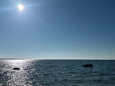 A bright sun illuminates a calm sea under a clear blue sky. The sun is positioned in the upper left quadrant of the image, casting a shimmering reflection across the water's surface. Two large rocks break the water in the foreground, one nearer to the viewer and the other further out. A small bird is perched on the further rock. On the horizon, a distant landmass is visible, with a faint outline of structures. In the far distance, a large ship can be seen on the water. The scene appears to be a coastal view from Tabasalu alevik, Estonia.