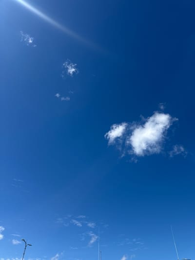 The image shows a clear blue sky with scattered white clouds. In the upper left corner, a bright streak of light, possibly from the sun, cuts across the sky. There are several small clouds dispersed across the sky, with a larger, more defined cloud formation in the middle right. The bottom of the image shows the tops of two streetlights and what appear to be thin antennas or lightning rods, suggesting an outdoor urban or suburban environment. The location is identified as Tiskre, Estonia, implying this is a view of the sky from that specific area. There are no people or animals visible in the image.