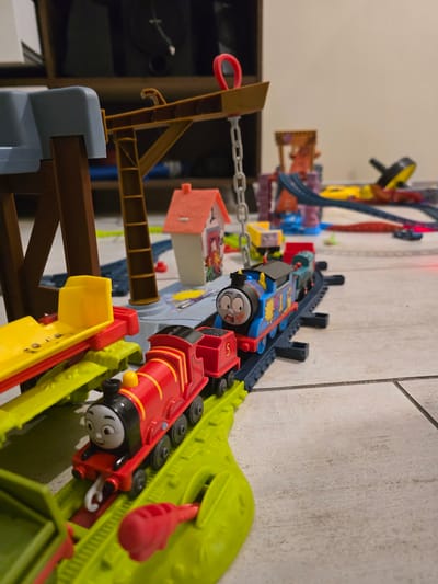 The image depicts a Thomas the Tank Engine train set. A red Thomas train is in the foreground attached to green and yellow components. Behind it is a blue Thomas train. The trains are on sections of toy tracks that are connected and integrated into a larger playset. The scene is situated on a tiled floor near a wall. There are no people present. The location is not identifiable from the image.