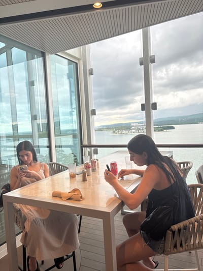 The image displays two young adult females seated at a square table with a light marble-like top and metal frame, located within a modern, glass-enclosed dining or lounge area. This area overlooks Falmouth Bay in Falmouth, Jamaica, offering a wide view of water, a distant landmass featuring green vegetation and scattered buildings, under an overcast sky.

The female on the left, with long dark hair, wears a light-colored sleeveless dress and operates a silver-backed smartphone with both hands. She is seated on a high-back woven bar stool.

The female on the right, also with long dark hair, wears a dark sleeveless top and dark shorts. She is similarly engaged with a black smartphone, holding it with both hands. A dark bag is draped over the back of her matching woven bar stool.

Centrally placed on the table are two empty clear glasses with ice residue, three soda cans (two red Coca-Cola, one white Coca-Cola Zero/Diet Coke), a small metallic shaker, and a folded beige cloth napkin.

The interior environment features tall glass panels secured by metal clamps to vertical white posts, a white slatted ceiling with a single recessed light fixture, and a dark floor. A white horizontal railing is visible beyond the table, separating the interior from the exterior view of the bay.