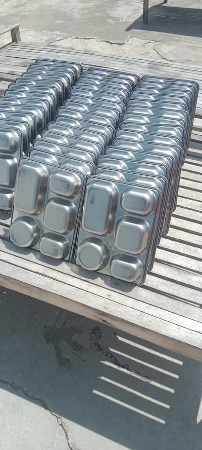 The image shows multiple stacks of metal trays, likely meal trays, arranged on a wooden surface. The trays are rectangular with divided compartments. The wooden surface appears to be a slatted bench or platform, which is situated on a concrete ground. Shadows from an unseen structure are cast across the ground. The environment suggests an outdoor setting, possibly a storage area or a canteen. The image is taken from a slightly elevated angle looking down. There are no people or animals visible in the image. The location is stated to be Puloblang, Indonesia, but this information is not visually verifiable from the image itself.