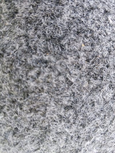The image displays a close-up, abstract view of a grey, textured surface with a fuzzy, fibrous appearance. The texture is composed of numerous fine, intertwined strands, creating a sense of depth and slight unevenness. There are subtle variations in shade, with some areas appearing lighter and others darker, suggesting a mottled or woven pattern. A few very small, indistinct yellow or brown specks are visible within the grey matrix. The overall impression is one of a soft, somewhat dense material, possibly fabric or a similar synthetic or natural fiber. There are no discernible people, objects, or activities depicted. The image provides no information about its location in the city of Shurchi, Uzbekistan, or any environmental context.