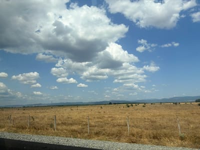 This is a wide-angle, outdoor, daytime photograph taken from a moving vehicle in the village of Zagortsi, Bulgaria. The scene depicts a dry, golden field bordered by a gravel road and a black asphalt surface in the foreground. A simple wire fence with wooden posts runs horizontally across the field. In the distance, a line of hills or mountains is visible under a bright blue sky filled with scattered white and grey clouds. The overall impression is of a rural landscape under a sunny, possibly hot, summer day. There are no people or animals visible in the image.