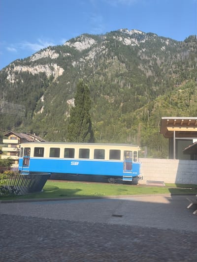 A blue and beige train car is parked on a track in Predazzo, Italy. The train car has multiple windows and the word "SSIF" is visible on its side. Behind the train, a mountain covered in trees rises against a clear blue sky. In the foreground, a paved area with a stone pattern is visible. A building with a wooden roof and a stone facade is located to the right of the train.