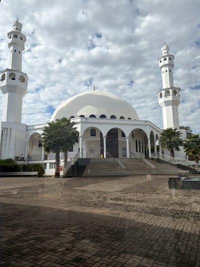 The image depicts the exterior of a white mosque with a large dome and two minarets. The mosque is situated in a paved courtyard. Palm trees are visible around the building, suggesting a warm climate. The sky is partly cloudy, indicating it could be daytime, possibly late morning or early afternoon, given the lighting and shadows. The architecture suggests a modern or contemporary design for the mosque. There are stairs leading up to the mosque entrance, with metal railings. The paving stones in the courtyard are laid in a patterned arrangement. Reflections are visible on the surface of the building, suggesting it might be made of polished material or there is glass in front of it. No people are visible in the scene. No text is legible in the image. The location context is Foz Do Iguaçu, Brazil.