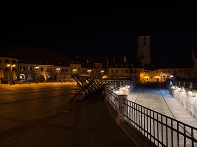 The image depicts a nighttime scene in Sibiu, Romania, specifically featuring the Bridge of Lies (Podul Minciunilor) connecting Piața Mare (Great Square) and Piața Mică (Small Square), with the Council Tower (Turnul Sfatului) visible in the background.
The foreground on the left shows a section of Piața Mare, an empty cobblestone square illuminated by several yellowish streetlights and distant building lights. Folded patio umbrellas are visible along the left edge. To the right, a black metal railing borders the descent of the Bridge of Lies, with three wall-mounted lamps illuminating a textured brick wall. The bridge itself features a prominent wooden support structure on its left side. Beyond the bridge, a cobblestone pathway descends into Piața Mică. The background is dominated by historic buildings with warm-toned facades and dark roofs. The Council Tower, a light-colored, multi-tiered structure with a visible clock face, stands illuminated in the center-right background, flanked by buildings with arched ground-floor passages. The sky is dark and clear. No people are present in the visible area.