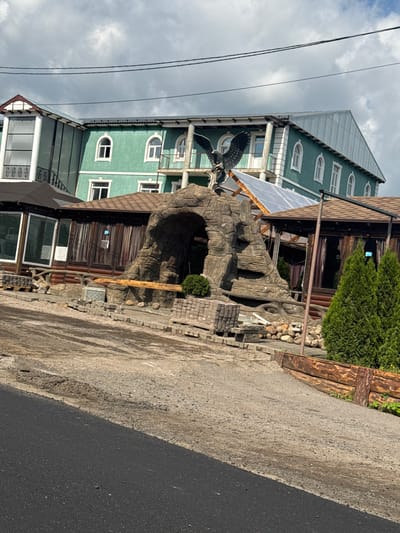 The image depicts a stone sculpture of an eagle with outstretched wings perched atop a faux rock formation. This sculpture is situated in front of a green, multi-story building with arched windows and a balcony. To the left and right of the rock formation are wooden structures with large windows and brown curtains, likely part of an establishment. The ground in front of these buildings is a mix of gravel and newly paved asphalt. Two coniferous trees stand to the right of the scene. The sky above is cloudy. The location is identified as Gorodok, Russia.