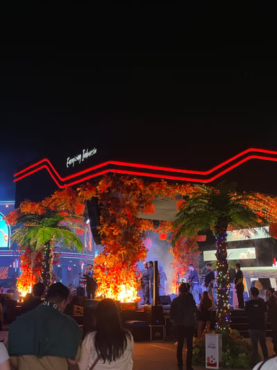 The scene shows an outdoor event, possibly a concert or festival, at night in Kosambi Barat, Indonesia. A stage is visible with performers and instruments, including a band with a guitarist and a keyboardist. The stage and surrounding area are decorated with trees and orange foliage, illuminated by string lights. A backdrop screen is present. Some audience members are visible in the foreground. A large sign with red neon-like lines and the words "Energizing Indonesia" is to the left. A "Fresh Cola" branded box is on the right. The lighting is low and atmospheric with a warm color cast, with some areas more brightly lit than others. Smoke or haze is present around the stage. The general atmosphere appears festive and lively.