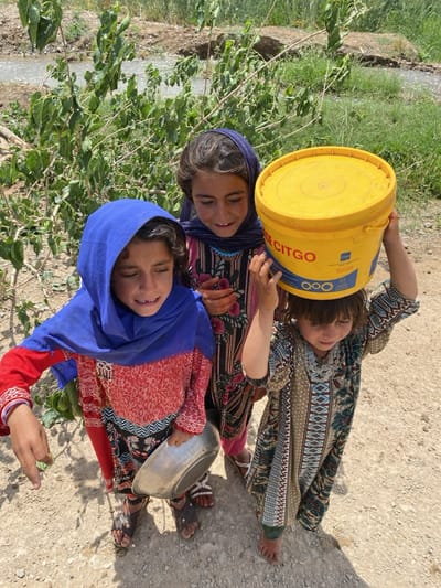 This outdoor vertical shot captures three young girls in Ashoqah, Afghanistan, standing on a dry, unpaved path under bright daylight. Lush green foliage and a small stream or canal with murky water are visible in the background.
On the far left, a girl wears a blue headscarf and a red, black, and white patterned dress, holding a silver metal bowl in her left hand. She has dark sandals on her feet. Beside her, a girl with a dark headscarf and a dark, patterned dress with floral motifs smiles, wearing a light blue beaded necklace.
The youngest girl is on the right, balancing a bright yellow bucket with a lid on her head, demonstrating focus. She is dressed in a long, flowing garment patterned with shades of green, brown, and blue, and is barefoot. The yellow bucket prominently features the word "CITGO" in blue text, along with "LUBE" and "BASE," and a circular gear-like logo.
The clear sunlight indicates it is daytime, casting visible shadows. The scene portrays a casual, everyday moment, possibly involving the collection or transport of water, reflecting rural life in Afghanistan. The expressions of the girls convey a sense of childhood and routine.