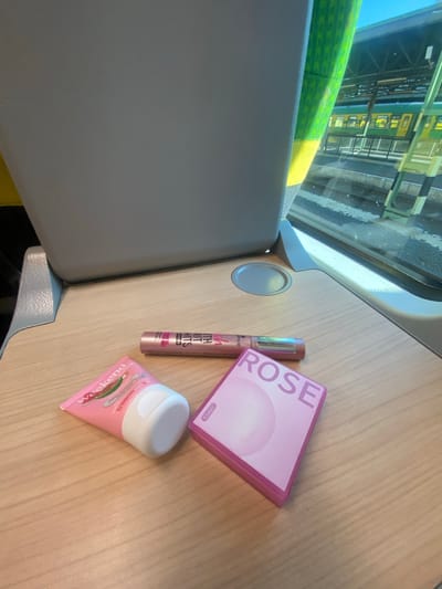 An overhead shot is captured of a tray table on a train with several cosmetic items placed upon it. In the foreground, a pink tube of "Weekend" hand cream is positioned to the left, featuring a picture of a watermelon slice. To its right, a tube of mascara with a holographic shimmer effect is lying horizontally. Further to the right, a pink square compact with "ROSE" printed in large letters is visible. In the background, the back of a grey train seat is seen, and through the window, a green and yellow train is visible at a station. The scene is likely taking place on a train traveling through or to Sopron, Hungary, given the context provided.