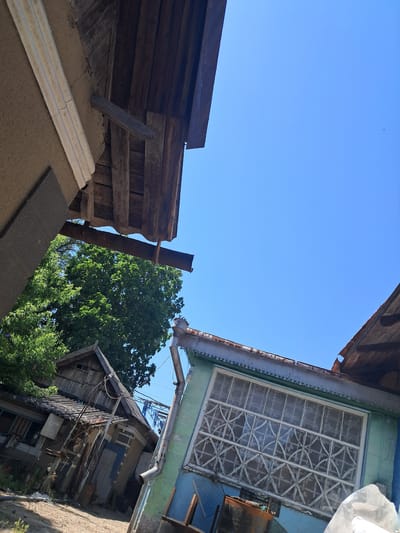 The image shows a low-angle view of the exterior of two dilapidated buildings under a clear blue sky. The building on the left appears to be made of wood and has a weathered, dark brown roof. A section of a beige-colored wall with a white trim runs along the left edge of the frame. The building on the right is painted a light teal color and features a large, multi-paned window with decorative white grilles. A rusty metal beam is visible extending from the roofline of the left building towards the right. Lush green foliage from a tree is visible between the two structures. The ground in the foreground is a sandy, uneven surface. The scene appears to be an outdoor area in a state of disrepair, possibly in a rural or semi-urban setting. The prompt specifies the location as Chobruchi, Moldova, though there are no visual cues within the image to confirm this.