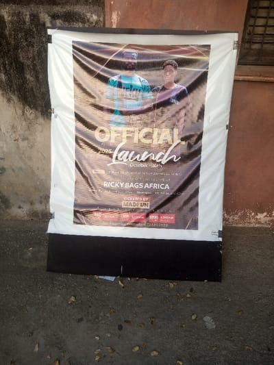 A vertical, wrinkled vinyl banner, advertising the "OFFICIAL Launch" of "RICKY BAGS AFRICA" for October 24, 2025, is displayed outdoors in Makadara/Mji Wa Kale ward, Kenya. The banner features two young adult males: one with a beard wearing a light blue/green patterned shirt, and another wearing glasses and a dark t-shirt. Event details include "TICKETED BY MADFUN" and ticket prices: REG 300Ksh, VIP 1,000Ksh, and ANNUL 4,000Ksh. A contact number, 0721857933, is provided for inquiries. The banner is affixed to a white frame, supported by a black base, and positioned against a light brown/orange, textured wall. The wall shows signs of wear and grayish-green discoloration on the left, with a small window or grate visible on the upper right. The ground in the foreground is dark grey, possibly concrete, with scattered brown leaves and white debris.