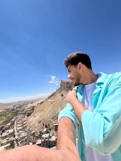 A man with dark hair and a beard, wearing a light blue shirt over a white t-shirt, is standing on a rocky outcrop overlooking the city of Ma'loula, Syria. He is looking down towards the city, which is nestled in a valley below a rocky landscape. The sky is clear and blue. The city consists of buildings with various rooftops. The ground is arid.