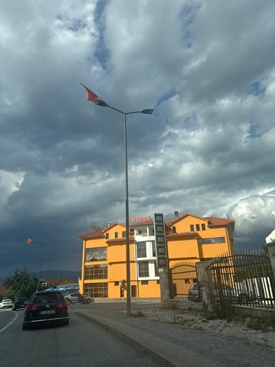 In the city of Resnik, Montenegro, a street-level, eye-level shot captures a cloudy sky over an orange building and a street. The building, featuring a prominent orange facade with large windows and a tiled roof, is partially obscured by a street lamp with a small red and yellow flag attached to it. The street in the foreground shows several cars, including a black Volkswagen Passat with a license plate reading "F AA 4877." A black fence with metal spikes borders a property to the right of the building. In the background, the dark storm clouds suggest approaching rain. The scene appears to be a typical urban streetscape with a mix of commercial and residential architecture under a dramatic sky.