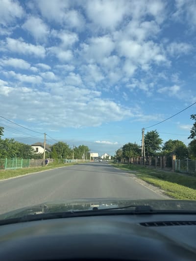 The image depicts a straight, paved road in the city of Velyki Hai, Ukraine. The road is lined with green grass on both sides, with fences and trees visible along the periphery. Several houses are nestled behind the fences, some with brick and some with wooden fencing. A single person is walking along the sidewalk on the left side of the road. Overhead, a bright blue sky is dotted with scattered white clouds. Power lines are strung across the upper portion of the image, supported by utility poles. The foreground shows the dashboard and windshield of a vehicle. The scene appears to be a typical daytime view of a residential street in a Ukrainian city.