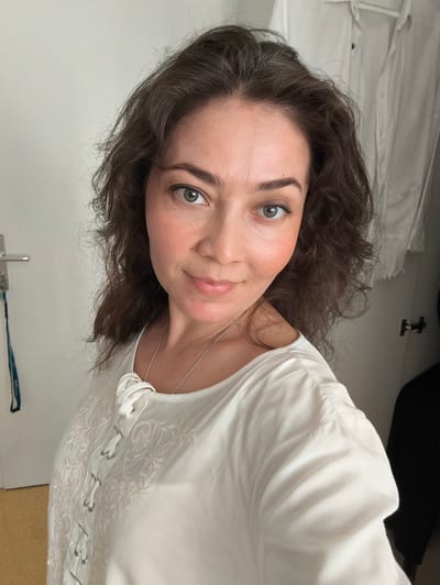 This is a medium shot selfie of a woman with shoulder-length, wavy brown hair. She has light skin with a subtle blush on her cheeks and green eyes. She is wearing a white, loose-fitting top with intricate embroidery and a lace-up detail at the neckline. A thin silver chain necklace is visible.
The woman is indoors, with a white wall behind her. To her left, a silver door handle is visible, and a blue lanyard is hanging from it. To her right, a white shirt is hanging on a hanger, and a dark object, possibly clothing, is draped over a closet door. The lighting suggests it is daytime. The woman is looking directly at the camera with a slight smile, conveying a sense of warmth and approachability. The setting appears to be a domestic environment, possibly a bedroom or dressing area, in Erlangen, Germany. There is no visible text in the image.