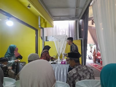 This media file captures an indoor or semi-outdoor social gathering in Kecamatan Gempol, Indonesia, likely during the daytime. The scene features several people, primarily adults, engaged in what appears to be a meal service or reception.
The setting includes vibrant yellow walls on one side, and on the other, white draped fabrics and a grey metal structure suggesting a temporary canopy or event space. Natural light filters in from an open area in the background, where a red carpet is visible, leading towards other guests. An artificial bright light bulb illuminates part of the yellow wall.
Several individuals are prominent. A young man, wearing a black batik shirt, stands near a table covered with a white tablecloth, holding a small white serving bowl, indicating he might be serving food or drinks. On the same table, a tissue box with "TISSUE" and "CLEAN" visible on its purple packaging is present. Further left, a woman in a green hijab and another in a black hijab are also near the food area, seemingly preparing or receiving items.
In the foreground, the backs of seated guests are visible. One man wears a traditional black peci (cap) and a patterned batik shirt. Another individual in a lighter-colored head covering is also seated. The attire, especially the batik shirts and hijabs, is common in Indonesian cultural or religious gatherings. The overall atmosphere suggests a communal event where people are mingling and partaking in refreshments.