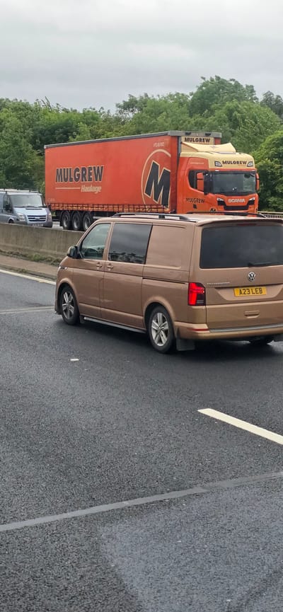 An orange semi-trailer truck with "MULGREW Haulage" written on the side is parked on a highway. The truck's cabin is yellow and orange, with "MULGREW" and "SCANIA" logos visible. A bronze Volkswagen Transporter van with license plate "A23 LEB" is in the foreground, also on the highway. In the background, a silver van with license plate "JF09 SXC" is visible. The scene appears to be a highway in Sheffield, United Kingdom, with trees lining the background. The sky is overcast.