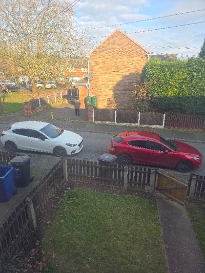 The image presents an elevated view of a residential street in Lichfield, United Kingdom, during daytime with partial cloud cover. Two hatchback automobiles are parked on the asphalt roadway: a white vehicle is positioned on the left, facing left, and a red vehicle is further back on the right, facing right. Both cars are equipped with dark alloy wheels. A single individual, dressed in dark clothing, is partially visible on a paved driveway behind the white car, located between a low brick wall and two green refuse bins; the person appears to be standing or walking away from the camera. In the foreground, a wooden picket fence with an open gate encloses a lawn area. Two refuse bins, one blue and one black, are situated by the street near the foreground fence. A prominent two-story brick building with a gable end is visible on the right side of the image, casting shadows onto the street. A large, bare deciduous tree occupies the left background, while dense green shrubbery borders the right side of the brick building. Overhead utility lines are visible against the sky. Fallen leaves are scattered on the ground, indicating an autumn or winter season.