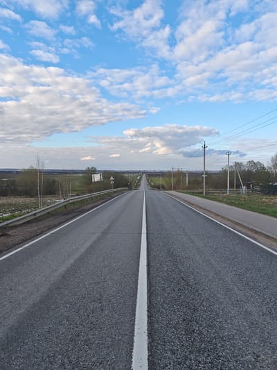 This image presents a wide view of a long, straight asphalt road extending into the distance under a vast sky, likely in Novosinkovo, Russia. The road, featuring a prominent white dividing line down its center and a solid white line along its right shoulder, appears to be a rural or intercity thoroughfare. The asphalt surface is dark grey with a visible gravel texture.
On the left side of the road, a metal guardrail runs parallel to the pavement. Beyond it, the landscape consists of green fields, some patches of lighter ground cover (possibly residual snow or dry grass), and several bare-branched trees, notably slender birches, and bushes. The terrain recedes into a flat, distant horizon.
To the right of the road, a narrow paved path or sidewalk runs alongside. Numerous utility poles with overhead power lines stretch into the horizon, mirroring the road's path. Further down, there are clusters of trees and a small, dark structure.
The sky is a bright blue, filled with scattered white, fluffy cumulus clouds, becoming denser towards the lower part of the sky. A faint crescent moon is discernible in the upper-left sky quadrant.
The time of day appears to be late afternoon or early evening, given the soft, diffused light and the visible moon. The weather is clear with moderate cloud cover. Several road signs are visible, including one on the left near the guardrail and others attached to utility poles on the right, but any text on them is illegible due to distance. The scene is devoid of people or explicit activity, conveying a sense of open space and tranquility.