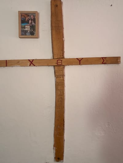 A wooden cross is affixed to a textured white wall. The cross is made of two rough-hewn wooden beams joined at their centers. The vertical beam is longer than the horizontal beam. Red markings are painted on the vertical beam, forming shapes that resemble the letters "M" and "Y" at the top, and an "X" and a horizontal line at the bottom. The horizontal beam has carved text that is difficult to discern clearly, but appears to be in Latin letters. To the left of the cross, a small framed photograph is hung on the wall. The photograph shows several people, some of whom are smiling. The scene appears to be indoors, given the wall and lighting. The overall impression is one of personal significance and remembrance.