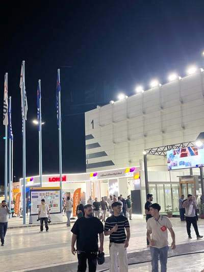 The image depicts an outdoor nighttime scene at an event, possibly an exhibition or festival, in Al-Ghizlaniya, Syria. In the background, a large, modern building with a white facade is illuminated by rows of bright lights. The building has a large number "1" prominently displayed on its upper section and appears to be divided into sections by dark horizontal bands. In front of this building, a large screen displays colorful images. Several flags on tall poles are visible on the left side of the frame, silhouetted against the dark night sky. The lower part of the image shows a paved area with many people walking around. Several stalls or booths are set up, with illuminated signage. One sign reads "LONGI" in large red letters. Another sign has a map-like display and the word "BUILDEX." Some people are walking, others appear to be talking or looking at displays. The overall atmosphere is one of a bustling public gathering at night.