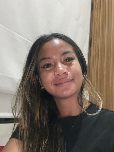 A woman is seen indoors, likely in a building in Tumbak Bayuh, Indonesia, although specific landmarks are not identifiable in the image. She is facing the camera with her head slightly tilted. She has dark hair with lighter highlights and is wearing a black shirt. The background includes a white wall and a wooden-patterned surface. The lighting is relatively bright.