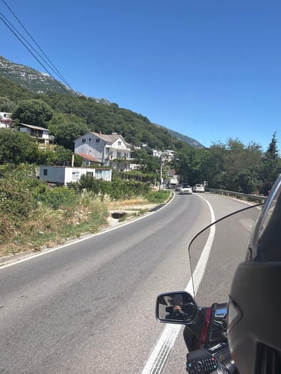 The image captures a view from a motorcycle traveling on a paved road in Mišići, Montenegro. The road curves to the right, with a white line marking its center and edge. On the left side of the road, there is a steep, tree-covered hillside with several white houses nestled among the foliage. The houses appear to be residential, with some featuring balconies and red-tiled roofs. On the right side of the road, there is a metal guardrail and more trees. In the distance, mountains rise, their peaks visible against a clear blue sky. Several cars are visible on the road ahead, including a white sedan and a white van. The foreground shows part of the motorcycle, including a windshield, a rearview mirror reflecting the rider's gloved hand and arm, and a control panel. The environment appears to be a sunny day in a rural or semi-rural area with a Mediterranean climate, given the lush greenery and the style of the houses.