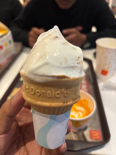 A person holds a McDonald's ice cream cone, focus on the cone and hand. In the background is a McDonald's tray with a cup, and what appears to be another person at the table. The location is not specified, therefore it cannot be located in Bencongan Indah, Indonesia.