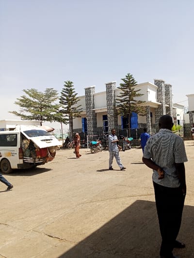 The image depicts an outdoor scene in the city of Toro, Nigeria, under a bright, clear sky. In the foreground, the back of a male subject wearing a grey patterned short-sleeve shirt and dark trousers is visible on the right, standing with his left hand positioned behind his back, casting a prominent shadow. The ground consists of compacted dirt and some concrete sections.
In the midground, a white passenger van, likely a commercial vehicle with "GOLALA" visible on its side, is parked on the left with its rear hatch open, revealing numerous bags and items packed tightly within and protruding from the back. To the right of the van, a female subject in a patterned dress walks rightward, slightly away from the viewer. A male subject wearing a light-colored plaid shirt and pants walks leftward in the central area. Further back, near the main building, multiple motorcycles are parked, including a light blue one and a darker one. Another male subject wearing a blue garment is partially visible further to the right.
The background features a modern multi-story building with light-colored walls and prominent grey, textured stone-clad pillars. Multiple rectangular openings, possibly windows or doors, are visible with blue coverings. Several trees are positioned in front of the building; two tall, slender evergreen trees and two broader deciduous trees with lighter green foliage.