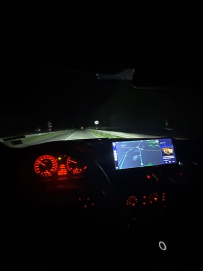 The image shows the interior of a vehicle driving at night. A navigation app is displayed on a smartphone mounted on the dashboard, showing a map with a route highlighted, potentially indicating the location in Mogila, Bulgaria. The vehicle's instrument cluster is illuminated in red, displaying gauges and indicators. The headlights illuminate the road ahead. The environment is dark, suggesting the vehicle is in an area with limited ambient lighting.