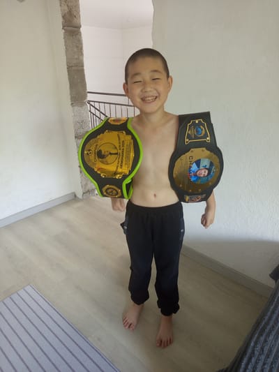 A young boy, smiling broadly, stands with two championship belts around his waist. The belts are black with gold detailing and brightly colored trim. The upper belt has a portrait of a man and text that reads "KAZAKHSTAN" and "CHAMPION". The lower belt has a green trim and features a circular emblem with a person's profile and the word "CHAMPION" in a golden arch. The boy is wearing black pants and is barefoot. He is in an indoor setting, with light-colored walls and a wooden floor. A section of a gray, ribbed blanket is visible in the upper right corner. The scene appears to be well-lit, suggesting daytime. The location is identified as Kenzheköl, Kazakhstan. The overall impression is one of pride and accomplishment. The text visible on the belts includes "KAZAKHSTAN", "CHAMPION", and Cyrillic text which appears to be related to the championship event and location.