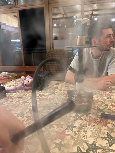 The image depicts an indoor scene, likely a residential area or a lounge, with a hookah prominently featured on a table covered with a patterned tablecloth. The table has a design of stars in red and white. A man is visible to the right, seated and looking away from the camera, with a cloud of smoke rising from the hookah. To the left, a plush purple monkey toy and a clear glass are placed on the table, along with a black handbag. The background shows wooden paneling and furniture, suggesting a rustic or cozy interior. The lighting is diffused, and the presence of smoke adds an atmospheric element to the scene. There is no specific indication of the city of Каменар, Bulgaria, within the visual information provided.