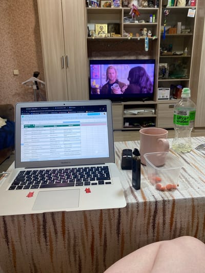 The image captures an indoor scene, likely in a home in Vyatskiye Polyany, Russia, featuring a person engaged in work or leisure on a laptop while a television plays in the background.

In the foreground, a silver MacBook Air laptop is open on a table, displaying a Google Sheets spreadsheet with content primarily in Russian. Visible text on the spreadsheet includes headings like "Список гостей" (Guest list), "Песня" (Song), and "Подсказки" (Hints), along with words such as "Свадьба" (Wedding), "Макая брат" (Makyai brother), and phrases like "работа не мужчина" (work is not a man). Two small red fox-like stickers adorn the laptop's palm rest.

Arranged on the table beside the laptop are several items: a green-capped plastic bottle of water labeled "Святой источник ultra+ water ВОССТАНОВЛЕНИЕ И ТОНУС" (Holy spring ultra+ water RECOVERY AND TONUS), a pink ceramic mug, a clear plastic container holding orange, spherical snacks, a black vape device, and a black remote control. A bare leg is partially visible on the far right, suggesting a person is seated at the table. The table itself has a textured surface with horizontal brown and beige stripes.

In the background, a television screen displays a scene with two individuals, one with dark hair and another with blonde hair holding a cup. To the left of the TV, a light-wood shelving unit is filled with various decorative objects, trinkets, and what appear to be books or files, contributing to a casual home atmosphere. Below the shelving unit are closed cabinets. The wall features patterned wallpaper. The overall impression is of an evening or night setting, given the indoor lighting and the TV being on.