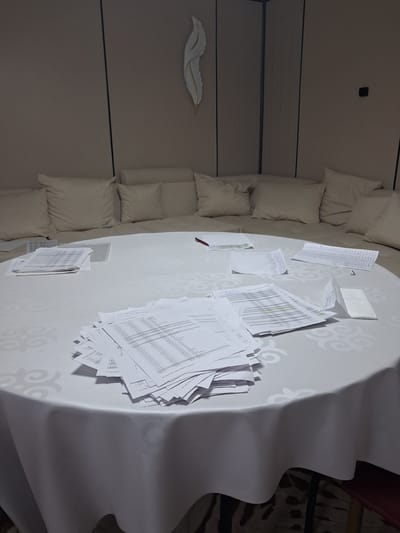 The image depicts a round table covered with a white tablecloth, laden with scattered stacks of papers, some neatly organized and others appearing disheveled. The papers seem to be documents with text and numerical data, suggesting a workspace or a meeting setting. The table is placed in a room with a beige upholstered sectional sofa in the background. A decorative feather-like object is mounted on the wall above the sofa. The lighting is subdued, casting a soft glow on the scene. The location is identified as Guldala, Kazakhstan. There are no individuals present in the image, and no discernible actions or interactions are taking place. The overall impression is one of a temporarily abandoned work area.