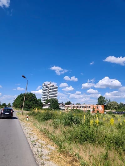 The image depicts a roadside scene under a bright blue sky with scattered white clouds. In the foreground, a black car is driving away from the camera on an asphalt road. The road is bordered by a grassy verge with dry grass and some green vegetation.

In the midground, there are two prominent buildings. The first is a tall, multi-story modern building with many windows, appearing to be in good condition. The second building is a longer, lower structure with an orange facade that shows signs of disrepair and weathering, with some windows broken or boarded up. Behind these buildings, there are various trees and foliage.

The scene is set in Krushovitsa, Bulgaria, as indicated by the metadata. The environment suggests a semi-urban or rural setting with a mix of functional and dilapidated structures. The presence of the road, car, and buildings points to an inhabited area. The clear sky and vegetation suggest a warm, likely summer day.