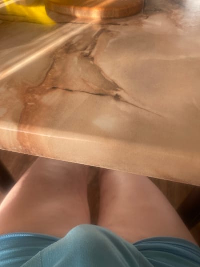 The image shows a close-up of a person's lap and legs, seen from a low angle, with a blurred countertop in the background. The person is wearing light blue shorts. The countertop appears to be made of a light-colored material with brown veining, and is illuminated by sunlight casting shadows. There is no discernible indication of the location being in Zhodzina, Belarus.