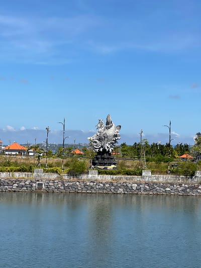 The image captures a view of the **Garuda Wisnu Kencana Cultural Park** in South Denpasar, Indonesia. The prominent subject is the partially visible, colossal statue of **Garuda**, a mythical bird-like creature, which is a key element of the larger Garuda Wisnu Kencana statue complex. The statue, rendered in a metallic, possibly stone or concrete, material, features intricate details of feathers and a dynamic posture. It is positioned on a raised platform with a concrete retaining wall and a base constructed from dark, rough-hewn rocks.
In the foreground, a body of calm, blue water reflects the sky and the surrounding environment. The water's surface shows slight ripples. Behind the statue and the rocky embankment, the park landscape unfolds. This includes patches of dry grass, scattered green shrubs and trees, and several ornamental lampposts with decorative elements.
Further in the background, there are structures with distinctive orange-tiled roofs, characteristic of Indonesian architecture. These buildings appear to be part of the park's facilities or surrounding areas. Beyond these, a hazy horizon shows more trees and buildings, suggesting an urban or semi-urban setting. The sky is a bright, clear blue with scattered, wispy white clouds. The overall scene is bathed in bright daylight, indicating a sunny day. There are no people visible in the image. The atmosphere is serene and monumental, highlighting the scale and artistic merit of the statue.