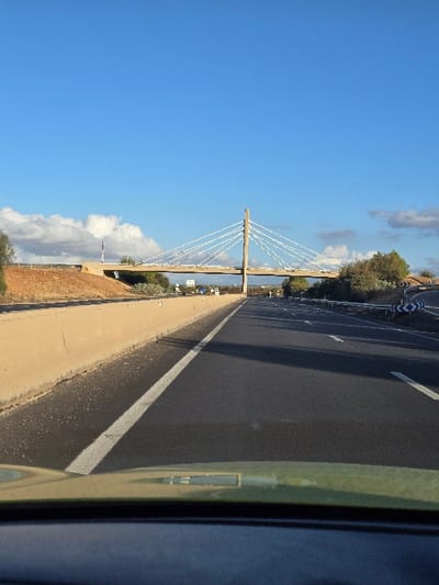 The image captures a bright, sunny daytime scene from the perspective of a vehicle traveling on a highway in Ain Cheggag, Morocco.

The highway occupies the right side of the frame, a dark asphalt surface with a distinct white line marking its edge, stretching into the distance. To the left of the road, there's a paved shoulder or walkway, bordered by a reddish-brown earthen embankment.

A prominent cable-stayed bridge is visible on the left side of the image, spanning horizontally across the middle ground. It features a tall, light-colored central pylon from which numerous white cables extend down to support the bridge deck. Beyond the bridge and along the horizon, green trees and vegetation are visible.

The sky above is a clear, vibrant blue with scattered white, fluffy clouds, suggesting excellent weather conditions. Part of the vehicle's frame, likely a door pillar or window edge, is visible along the far right edge, indicating the photo was taken through a car window. There is no discernible activity involving people, nor is any readable text visible on signs or objects within the frame.
