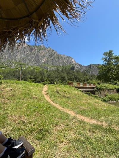 The image depicts a scenic outdoor environment with a prominent rocky mountain in the background. In the foreground, there is a patch of green grass with a dirt path winding through it. A portion of a thatched umbrella is visible in the upper left corner. Further in the background, a forest of green trees lines the base of the mountain. A bridge with red and white railings spans a body of water, and to the right of the bridge, there is a large green tree. A small part of a person's legs wearing black athletic shoes and white socks is visible in the lower left corner, seated on what appears to be a wooden bench. The sky is clear and blue. The location is identified as Zgorigrad, Bulgaria.