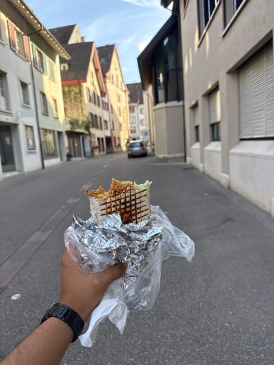 The image, correctly oriented, captures a person's hand holding a takeaway food item on a sunny street in Liestal, Switzerland. The central subject is a grilled wrap or sandwich, with visible char marks on the tortilla and some filling, including lettuce and other components, peeking out. It is partially encased in aluminum foil and a clear plastic bag. A black, possibly digital, wristwatch with a metal band is worn on the person's left wrist.
The scene unfolds in an outdoor urban setting, likely a street or alleyway. On the left, several traditional multi-story buildings line the street, showcasing varied facades in hues of yellow, brown, and off-white, characteristic of European architecture. Some feature pitched roofs and distinct window styles. On the right, the buildings appear more contemporary or commercial, with larger windows, some equipped with blinds, and a generally more uniform greyish-beige color palette. The street itself is paved with asphalt or a similar material.
The activity depicted is an individual holding a street food meal, suggesting they are about to eat or have just started. The bright, clear sky and distinct shadows indicate a sunny day, likely late morning or afternoon. No specific emotions are discernible, and no visible text can be read on the food packaging or buildings. The overall atmosphere is that of a pleasant, everyday moment in a Swiss town.