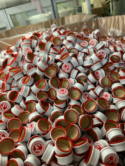 A pile of white bottle caps with red trim and red logos is shown. The bottle caps are made of metal and have a textured pattern around the side. The logos are circular and depict a stylized "S" in red. The bottle caps are piled in a large quantity, appearing to be in a warehouse or manufacturing facility. In the background, cardboard boxes and shelving units can be seen, suggesting this is a storage or processing area. The lighting is artificial and somewhat dim. The overall scene is one of bulk packaging materials.