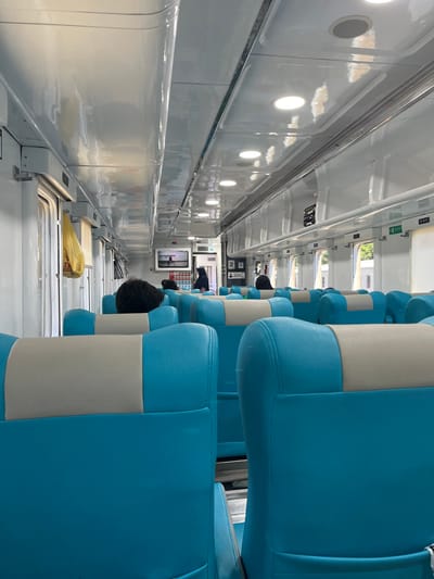 The image depicts the interior of a passenger train carriage, likely in Indonesia. The seats are upholstered in a combination of bright blue and beige. The carriage features an overhead luggage rack and fluorescent lighting. A yellow plastic bag is hanging from a hook near one of the windows. Several passengers are visible, some with their backs to the camera. The overall impression is of a clean and functional train interior. The location is specified as Desa Tambakreja, Indonesia.