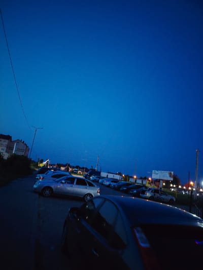 The image depicts a nighttime scene in a city, likely at dusk or early night. The sky is a deep, clear blue, indicative of twilight. A street lined with parked cars is visible, with a row of buildings on the left side of the frame. These buildings appear to be multi-story residential or commercial structures. Streetlights and lights from the buildings illuminate the scene. In the background, a billboard with Russian text is present, suggesting a commercial area. The overall atmosphere is calm and quiet, characteristic of a residential or suburban setting at night. It is impossible to determine the exact location of Крутые Выселки, Russia from the visual information provided.