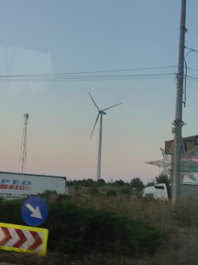 The image depicts a rural or industrial landscape, likely in or around Adunații-Copăceni, Romania, with a focus on renewable energy infrastructure and road signage. A prominent wind turbine stands in the middle ground, its white blades stark against a pale blue and pink sky, suggesting either dawn or dusk. To the left of the turbine is a tall, lattice-style telecommunications tower. In the foreground, a white semi-trailer truck with partially visible lettering, "PED," is parked. A blue circular sign with a white arrow pointing to the lower right and a yellow and red chevron-patterned barrier are visible in the lower left quadrant of the image, indicating a directional change or hazard. A utility pole with wires runs along the right side of the frame. A white truck cab is partially visible through overgrown vegetation in the middle right of the image. The scene is framed by dry grass and bushes. The overall impression is one of a functional landscape, possibly on the outskirts of a town or near a transport route.
