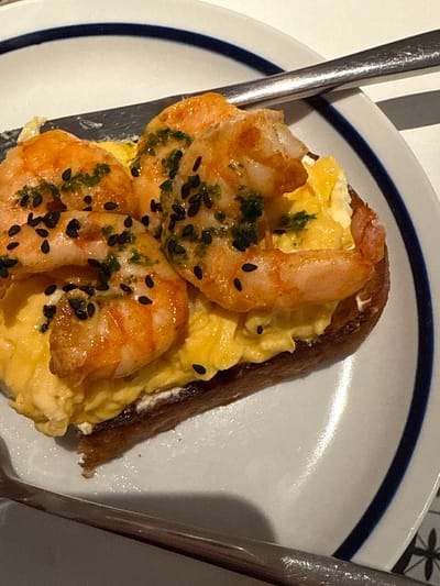 A close-up shot of a plate featuring a piece of toasted dark bread topped with scrambled eggs and three cooked shrimp. The shrimp are seasoned with black sesame seeds and green herbs. Two pieces of silverware flank the plate on either side. The plate is white with a blue rim. The location information provided is irrelevant to the visual content of the image.