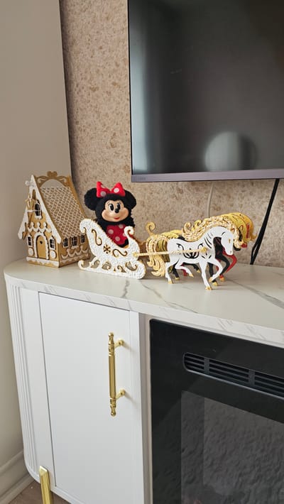 In Sof'ino, Russia, on a white marble-topped cabinet with a gold handle, a Minnie Mouse plush toy wearing a red bow and polka dot dress sits in a white and gold sleigh. The sleigh is pulled by three decorative horses, two white with gold manes and accents, and one black with red accents and a gold mane. To the left of the sleigh is a gold and white gingerbread house. Behind the display is a television screen and a textured wallpapered wall. The cabinet has white doors and a black fireplace insert.