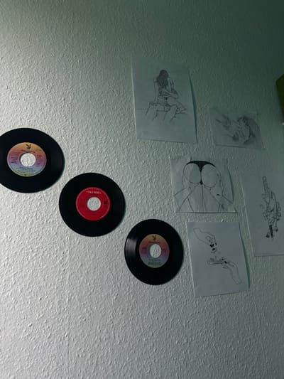 A collection of vinyl records and sketches are displayed on a textured, white wall. Three vinyl records, two black and one with a red label, are arranged on the left side of the frame. The records have labels with text and graphics, indicating they are singles. To the right of the records, several line-drawing sketches are affixed to the wall. These sketches depict various subjects, including human figures in intimate poses, a stylized anatomical drawing of buttocks, a hand rolling a cigarette, and a drawing of a mask. The lighting is dim, casting soft shadows on the wall and highlighting the textures. The scene appears to be in a domestic setting, possibly a bedroom or living area. The image does not contain any elements that would allow for determining its specific location within the city of Roskilde, Denmark.