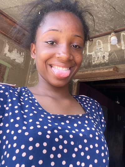 A young Black female is depicted in a close-up, chest-up portrait, likely a selfie, taken in Idiroko, Nigeria. She is smiling broadly with her tongue extended slightly from her mouth, exposing her upper teeth. Her hair is dark and appears loosely styled. She wears a dark blue top or dress adorned with numerous light-colored polka dots, featuring a rounded neckline. The background consists of a wall exhibiting signs of wear, including peeling paint or plaster, and rough texture. To the subject's left, faded dark markings or drawings are visible on the wall, one resembling a rudimentary structure. A light bulb in a white socket hangs from a wire in the upper right background. A wooden frame or beam runs horizontally behind the subject's head.