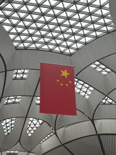 A low-angle shot captures the interior of a modern building with a triangular-paned glass ceiling. A large red flag with five yellow stars, the flag of the People's Republic of China, hangs vertically from a thin cord in the center of the frame. The ceiling is comprised of a geometric grid of triangles and the building's interior walls are white and curved, with black structural beams visible. The scene is brightly lit by natural light filtering through the ceiling.